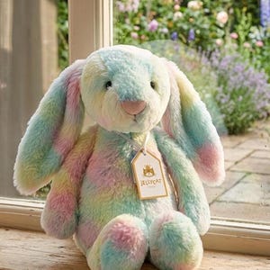 May include: A pastel-colored plush bunny with long floppy ears and a pink nose. The bunny has a tag that says "Jellycat Design". The bunny is sitting on a wooden surface in front of a window with a garden view.