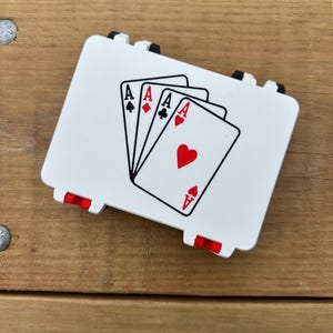 May include: A white playing card holder with four Aces displayed. The holder has black and red accents and is set against a wooden background. The Aces are in the suits of spades, diamonds, clubs, and hearts.