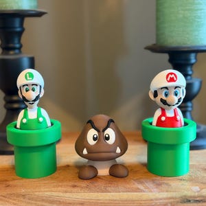 May include: Two plastic figurines of characters wearing white hats with red or green emblems, each emerging from a green cylindrical pipe. Between them sits a brown, mushroom-shaped character with a stern expression and two white fangs.