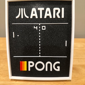 May include: A framed black and white Atari Pong game display. The Atari logo is at the top, with the word PONG at the bottom. The score is 4-0. The frame is white.