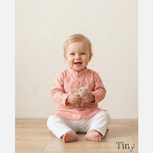 May include: A long-sleeved peach-coloured shirt with a subtle diamond-patterned eyelet texture, featuring a mandarin collar and three small gold buttons at the neckline, paired with plain white trousers and a small beige plush toy.