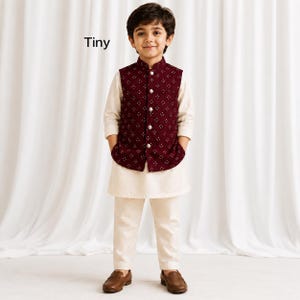 May include: A child wearing a cream-colored long-sleeved kurta and matching trousers paired with a dark maroon sleeveless vest featuring a white geometric diamond pattern and silver buttons. The word Tiny is printed in black text on the left side.