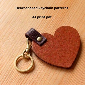 Peut inclure: Un porte-clés en cuir en forme de cœur avec un cœur en cuir marron, une lanière marron foncé et un fermoir doré. Le texte "Heart-shaped keychain patterns" et "A4 print pdf" sont visibles.