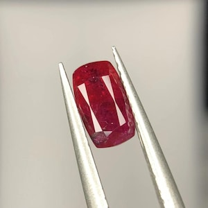 May include: A cushion-cut ruby gemstone with a deep red color and faceted surface held by a pair of silver-toned metal tweezers. The stone displays internal clarity characteristics and sharp geometric light reflections across its top surface.