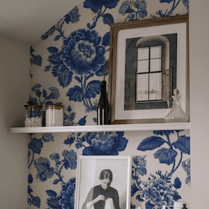 May include: A shelf with decorative items against a blue and white floral wallpaper. Items include framed art, glass jars, a dark bottle, and a clear glass decanter. A second framed picture is on the lower shelf.