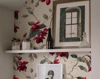 May include: A shelf displays a framed picture of a window, a black bottle, and a glass decanter. Below, a framed black and white photo of a person sewing. The background features floral wallpaper with red flowers and green leaves.
