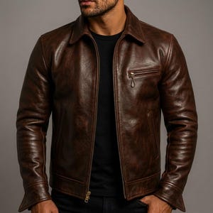 May include: A dark brown leather jacket with a classic collar, a full-length front zipper, and a single zippered chest pocket on the left side. The jacket features long sleeves with buttoned cuffs and a smooth, polished finish throughout the exterior.