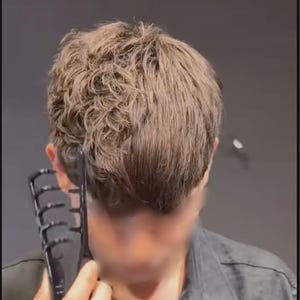 Puede incluir: Un peine negro se usa en cabello corto y castaño claro. El peine tiene dientes anchos y se sostiene cerca de la parte posterior de la cabeza. La cara de la persona está borrosa y lleva una camisa oscura.