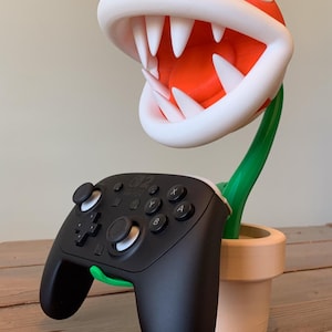 Puede incluir: Una planta Piraña impresa en 3D de la serie de videojuegos Super Mario, sosteniendo un mando Nintendo Switch Pro negro. La planta tiene una cabeza roja con dientes blancos y un tallo verde, en una maceta beige.