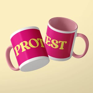 May include: Two ceramic mugs with a bright pink exterior and interior. The mug on the left features the yellow text PROT, while the mug on the right displays the yellow text EST. Both mugs have a white border around the text and a pink handle.