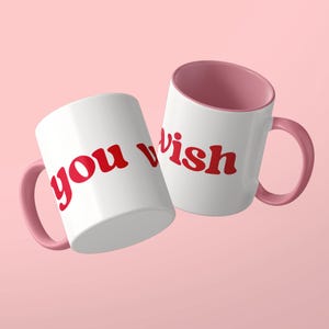 May include: Two white ceramic mugs with pink interiors and matching pink handles. The mugs feature the red text "you wish" printed in a bold, rounded, lowercase font. The mugs are positioned at an angle, displaying the text clearly on the white exterior surface.