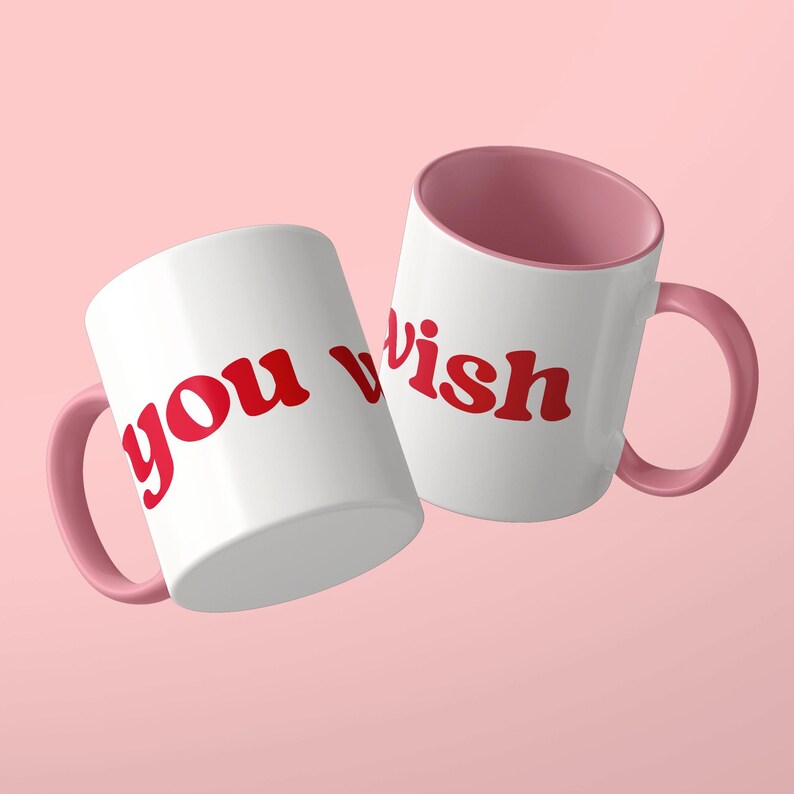 May include: Two white ceramic mugs with pink interiors and matching pink handles. The mugs feature the red text "you wish" printed in a bold, rounded, lowercase font. The mugs are positioned at an angle, displaying the text clearly on the white exterior surface.