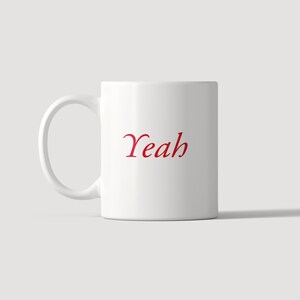 May include: A white ceramic mug featuring a handle on the left side and the word Yeah printed in a red serif font in the center. The mug has a smooth, cylindrical body and a clean, simple design, providing a clear view of the text on the front.