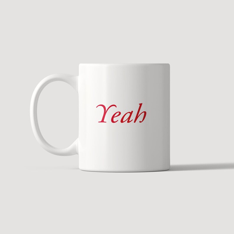 May include: A white ceramic mug featuring a handle on the left side and the word Yeah printed in a red serif font in the center. The mug has a smooth, cylindrical body and a clean, simple design, providing a clear view of the text on the front.