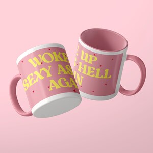Może przedstawiać: Dwa r&oacute;żowe ceramiczne kubki z ż&oacute;łtym napisem WOKE UP SEXY AS HELL AGAIN. Kubki mają zakrzywiony uchwyt i gładkie wykończenie, ozdobione małymi czerwonymi gwiazdkami rozrzuconymi na r&oacute;żowej powierzchni cylindrycznych naczyń.