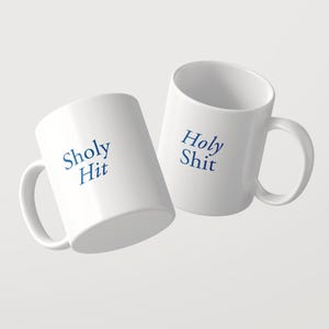 May include: Two white ceramic mugs with a glossy finish and a C-shaped handle. One mug features the blue text Sholy Hit, while the other displays the blue text Holy Shit. The mugs are positioned at an angle, showcasing the printed text clearly.