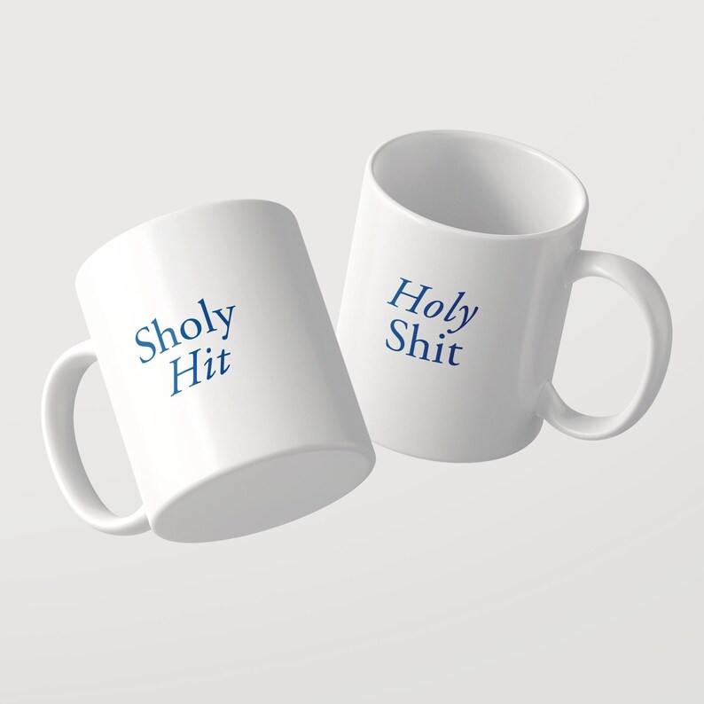 May include: Two white ceramic mugs with a glossy finish and a C-shaped handle. One mug features the blue text Sholy Hit, while the other displays the blue text Holy Shit. The mugs are positioned at an angle, showcasing the printed text clearly.