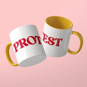 May include: Two white ceramic mugs with yellow handles and interiors. The left mug features the red text PROT, while the right mug displays the red text EST. The mugs are positioned at an angle against a solid light pink background.