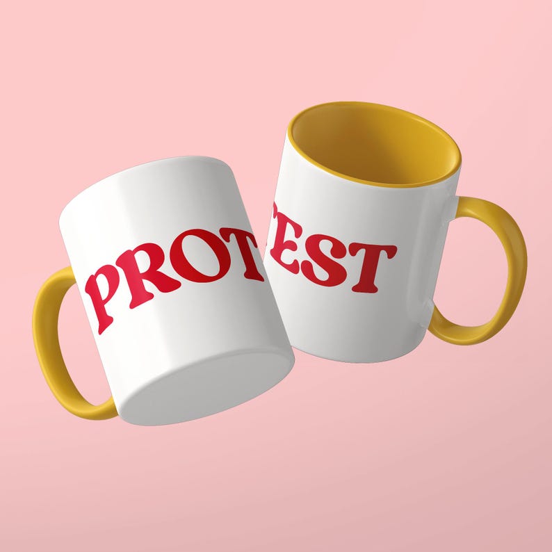 May include: Two white ceramic mugs with yellow handles and interiors. The left mug features the red text PROT, while the right mug displays the red text EST. The mugs are positioned at an angle against a solid light pink background.