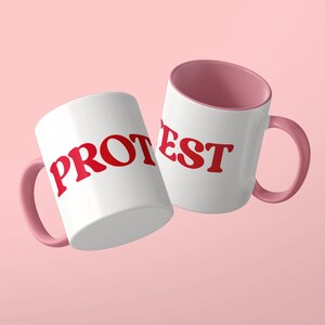 May include: Two white ceramic mugs with pink interiors and pink handles. The left mug features the red text PROT and the right mug features the red text EST. The mugs are positioned at an angle against a solid light pink background.