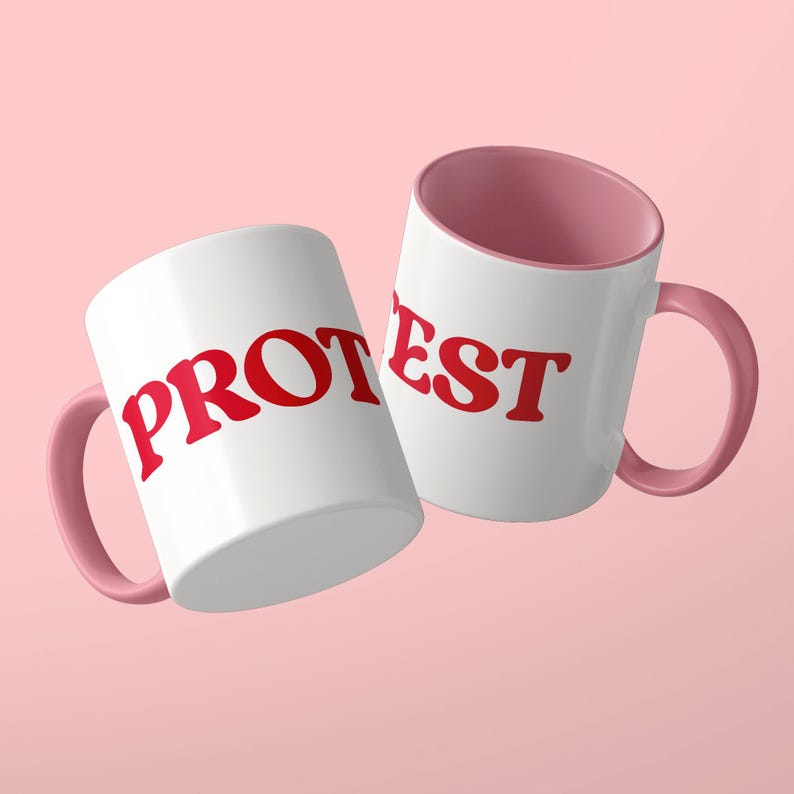 May include: Two white ceramic mugs with pink interiors and pink handles. The left mug features the red text PROT and the right mug features the red text EST. The mugs are positioned at an angle against a solid light pink background.