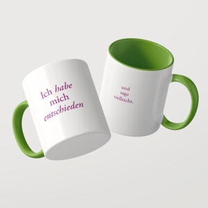 May include: Two white ceramic mugs with green interiors and green handles. The first mug features the purple text Ich habe mich entschieden, while the second mug displays the purple text und sage vielleicht. Both mugs have a smooth finish.