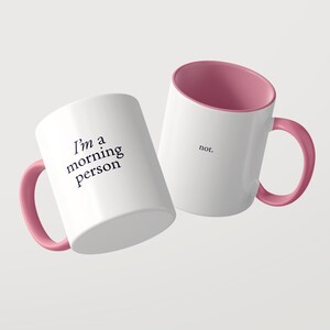 Może przedstawiać: Dwa białe ceramiczne kubki z r&oacute;żowym wnętrzem i uchwytem. Jeden kubek zawiera czarny napis 'I'm a morning person', a drugi kubek zawiera czarny napis 'not.' Kubki są ustawione pod kątem na jasnym, jednolitym tle.