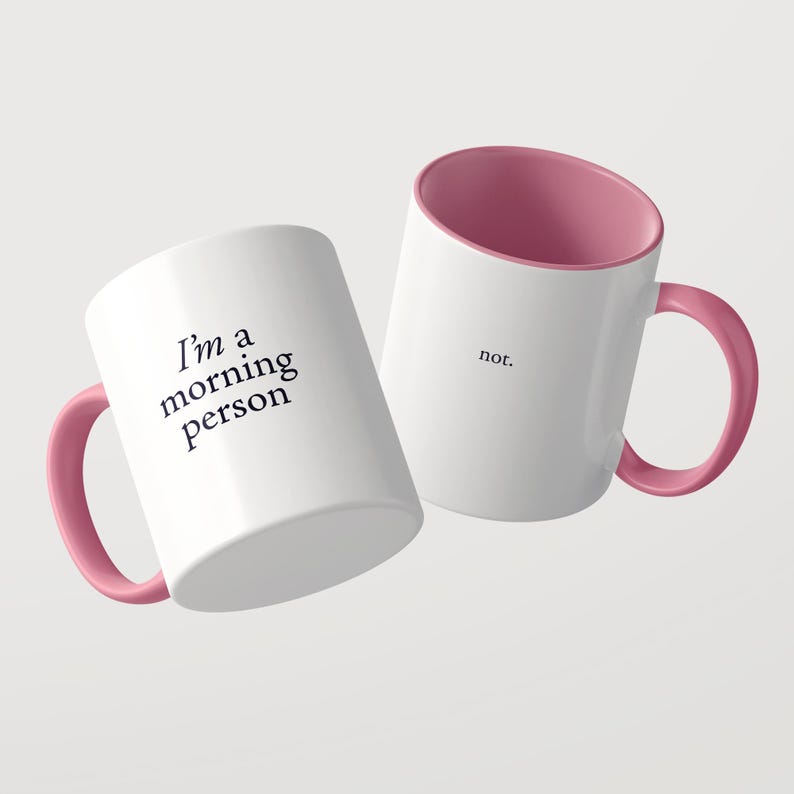 Może przedstawiać: Dwa białe ceramiczne kubki z r&oacute;żowym wnętrzem i uchwytem. Jeden kubek zawiera czarny napis 'I'm a morning person', a drugi kubek zawiera czarny napis 'not.' Kubki są ustawione pod kątem na jasnym, jednolitym tle.