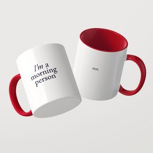 Może przedstawiać: Dwa białe kubki ceramiczne z jaskrawoczerwonym wnętrzem i uchwytem. Jeden kubek posiada czarny napis 'I'm a morning person', a drugi napis 'not.' Kubki są ustawione pod kątem na jasnym tle.