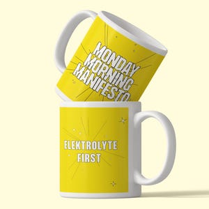 May include: Two white ceramic mugs with yellow exterior panels. The top mug features the black text MONDAY MORNING MANIFESTO with radiating lines. The bottom mug features the white text ELEKTROLYTE FIRST with radiating lines and small star icons.
