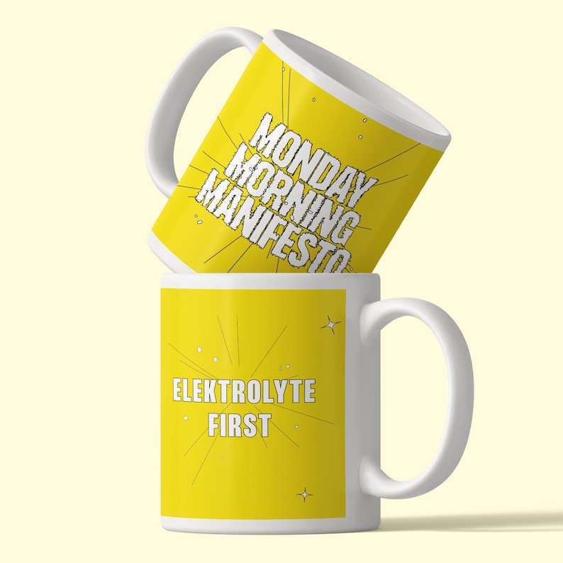 May include: Two white ceramic mugs with yellow exterior panels. The top mug features the black text MONDAY MORNING MANIFESTO with radiating lines. The bottom mug features the white text ELEKTROLYTE FIRST with radiating lines and small star icons.