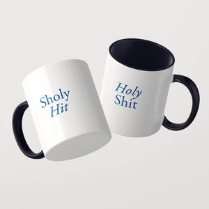 May include: Two white ceramic mugs with black interiors and black handles. One mug features the blue text Sholy Hit, while the other mug displays the blue text Holy Shit. The mugs are positioned at an angle against a plain light gray background.