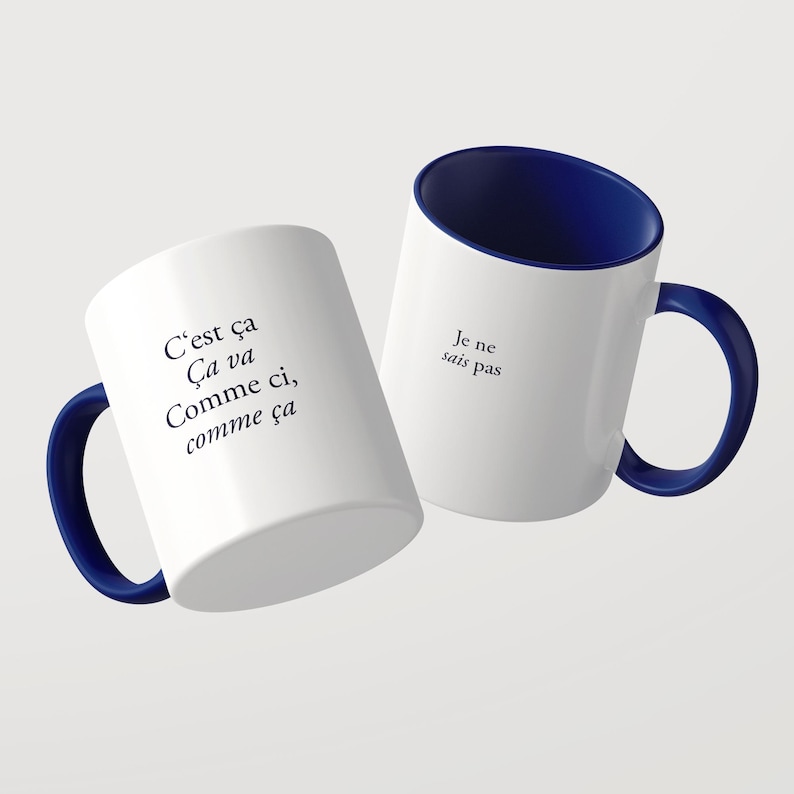 May include: Two white ceramic mugs with dark blue interiors and handles. One mug features the black text C'est &ccedil;a &Ccedil;a va Comme ci, comme &ccedil;a. The other mug displays the black text Je ne sais pas. Both mugs have a smooth finish and a cylindrical shape.