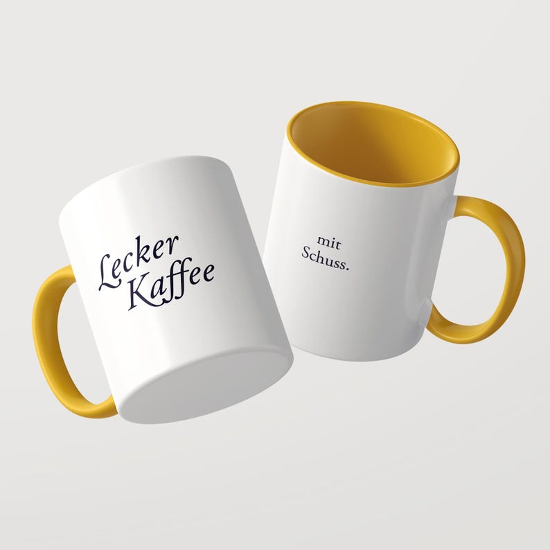 May include: Two white ceramic mugs with yellow interiors and yellow handles. One mug features the black text Lecker Kaffee, while the other mug displays the black text mit Schuss. The mugs are positioned at an angle against a plain background.
