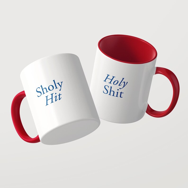 May include: Two white ceramic mugs with red interiors and red handles. One mug features the blue text Sholy Hit, while the other mug displays the blue text Holy Shit. The mugs are positioned at an angle against a plain light gray background.