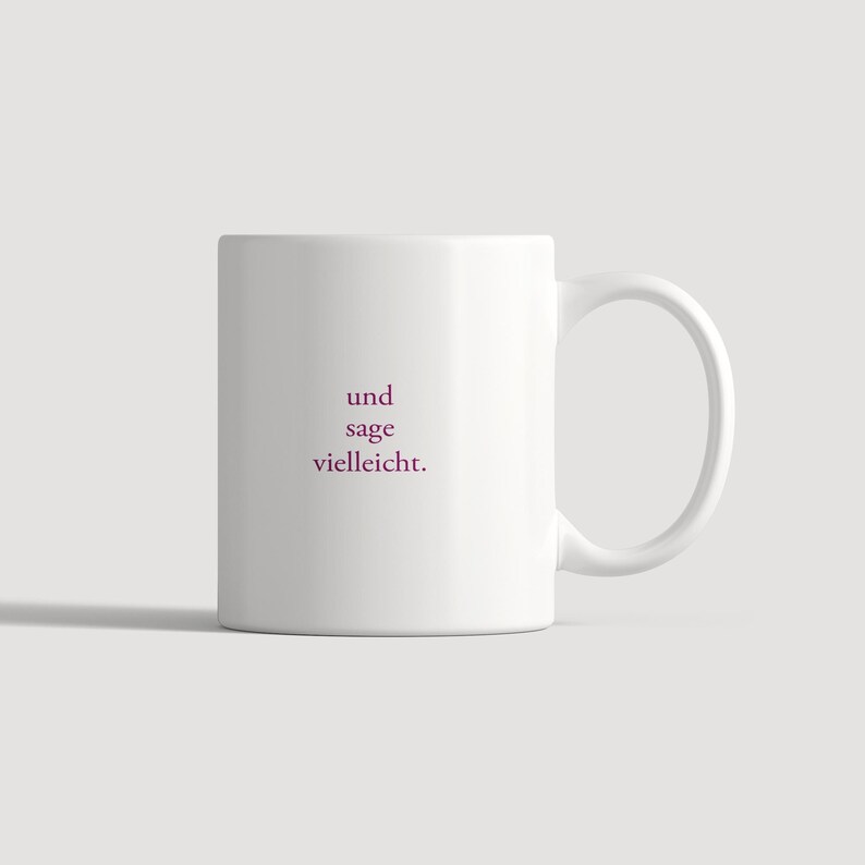 May include: A white ceramic mug featuring the dark pink text "und sage vielleicht." printed in the center. The mug has a smooth, cylindrical body and a curved handle on the right side, providing a simple and functional design for holding beverages.