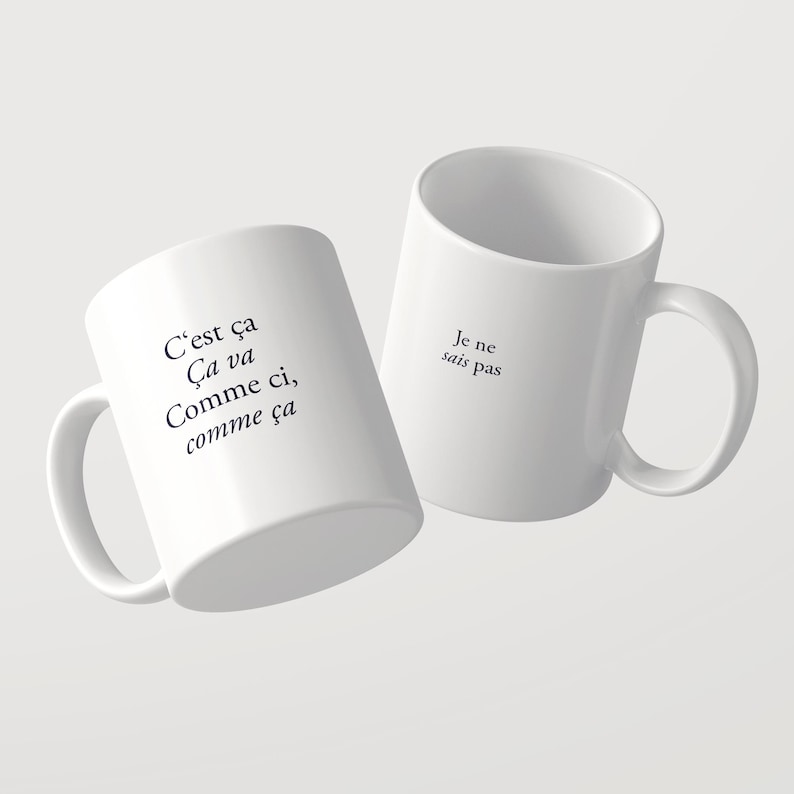 May include: Two white ceramic mugs featuring black printed text. One mug displays the French phrase C'est &ccedil;a &Ccedil;a va Comme ci, comme &ccedil;a, while the other mug features the phrase Je ne sais pas. Both mugs have a standard handle and cylindrical shape.