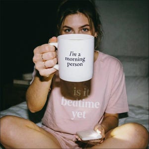 Może przedstawiać: Biały ceramiczny kubek z czarnym nadrukiem 'I'm a morning person' oraz jasnor&oacute;żowy t-shirt z białym nadrukiem 'is it bedtime yet' z przodu, noszony przez osobę trzymającą kubek i małe białe urządzenie w dłoniach.