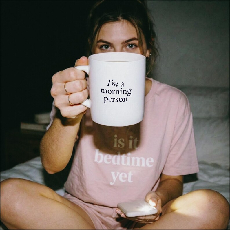 Może przedstawiać: Biały ceramiczny kubek z czarnym nadrukiem 'I'm a morning person' oraz jasnor&oacute;żowy t-shirt z białym nadrukiem 'is it bedtime yet' z przodu, noszony przez osobę trzymającą kubek i małe białe urządzenie w dłoniach.