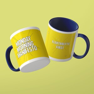 May include: Two ceramic mugs with dark blue interiors and handles. The exterior is yellow with white text. One mug features the text MONDAY MORNING MANIFESTO, and the other features the text ELEKTROLYTE FIRST, both surrounded by thin radiating lines.