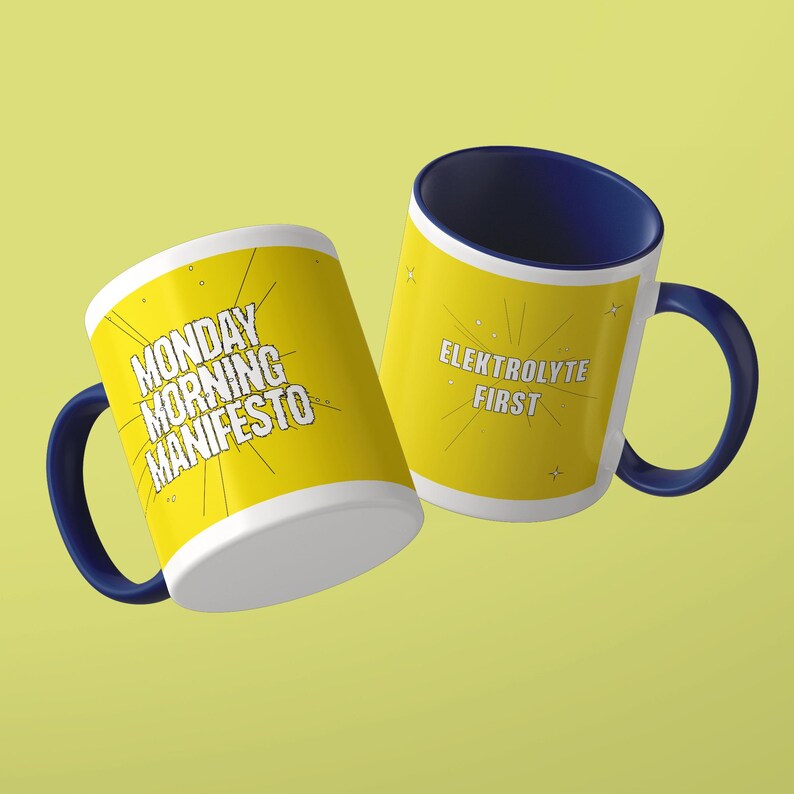 May include: Two ceramic mugs with dark blue interiors and handles. The exterior is yellow with white text. One mug features the text MONDAY MORNING MANIFESTO, and the other features the text ELEKTROLYTE FIRST, both surrounded by thin radiating lines.