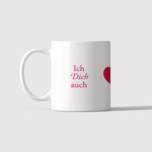 Pu&ograve; includere: Una tazza in ceramica bianca con il testo Ich Dich auch stampato in un carattere serif rosa scuro, accompagnato da una forma di cuore parziale rosa scuro sul lato destro. La tazza presenta un corpo cilindrico standard e un manico curvo a sinistra.
