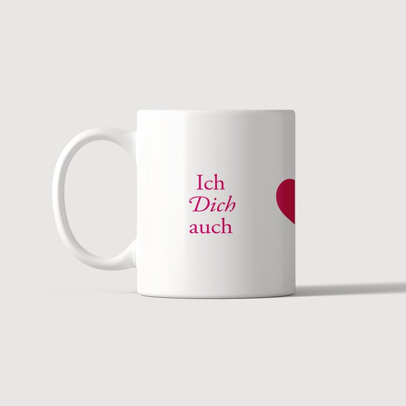 Pu&ograve; includere: Una tazza in ceramica bianca con il testo Ich Dich auch stampato in un carattere serif rosa scuro, accompagnato da una forma di cuore parziale rosa scuro sul lato destro. La tazza presenta un corpo cilindrico standard e un manico curvo a sinistra.