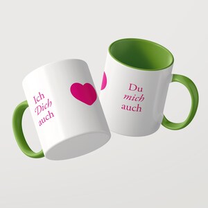 Pu&ograve; includere: Due tazze in ceramica bianca con interno e manico verdi. La tazza a sinistra presenta il testo magenta Ich Dich auch e un cuore magenta. La tazza a destra mostra il testo magenta Du mich auch. Entrambe le tazze hanno una finitura liscia.