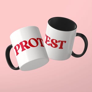 May include: Two white ceramic mugs with black handles and black interiors. The mugs feature the red text PROTEST printed in a bold, serif font across the exterior surface, with the letters split between the two mugs for visibility.