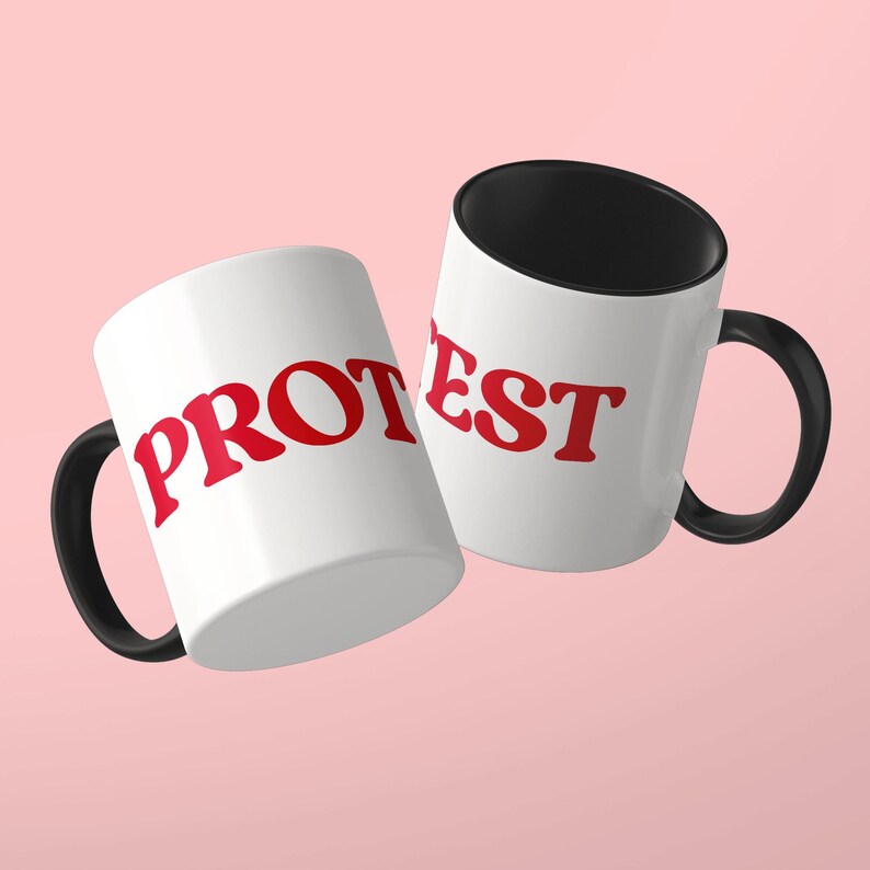 May include: Two white ceramic mugs with black handles and black interiors. The mugs feature the red text PROTEST printed in a bold, serif font across the exterior surface, with the letters split between the two mugs for visibility.