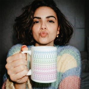 Puede incluir: Una taza de cer&aacute;mica blanca con l&iacute;neas de texto horizontales repetidas en azul, verde y morado. El texto dice 'up' en azul, 'the' en verde y 'sun' en morado, dispuesto en un patr&oacute;n apilado alrededor del exterior de la taza cil&iacute;ndrica.