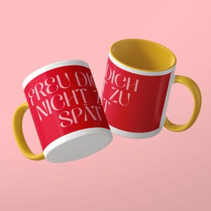 May include: Two ceramic mugs with a bright red exterior and a yellow interior and handle. The mugs feature white text that reads "FREU DICH NICHT ZU SP&Auml;T" in a stylized, elegant font. The design is consistent across both mugs, showing the front and side.