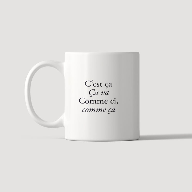 May include: A white ceramic mug with a handle on the left side. The front of the mug features black printed text that reads: C'est &ccedil;a, &Ccedil;a va, Comme ci, comme &ccedil;a. The mug has a smooth, cylindrical body and a clean, simple design.