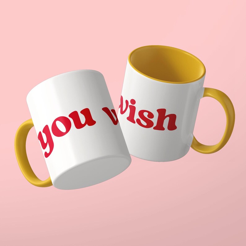 May include: Two white ceramic mugs with yellow interiors and yellow handles. The mugs feature the red text "you wish" printed in a bold, rounded font. The mugs are positioned at an angle against a solid light pink background.